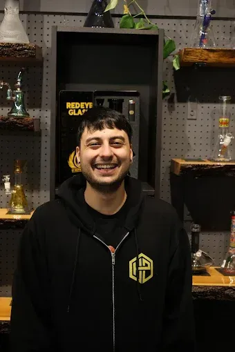A person with dark hair wearing a black zip-up hoodie smiles in front of a shop display of glass pipes and accessories in a Niagara Falls dispensary