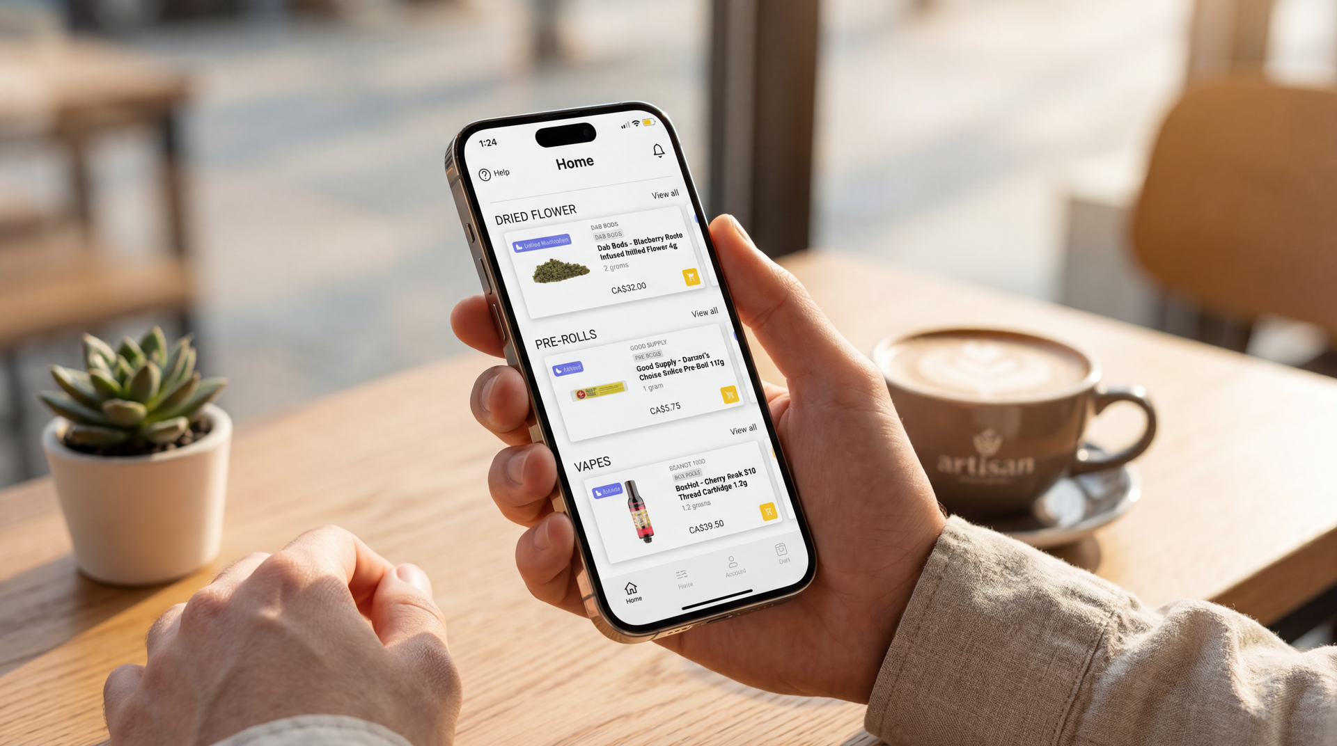 A person holds a smartphone displaying a shopping app, sitting at a wooden table with a coffee cup and a small plant.