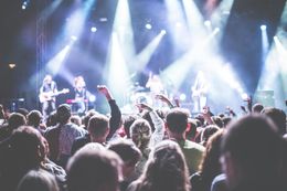 Concert crowd facing a band on stage, bright lights shining. People raise arms, enjoying the show.