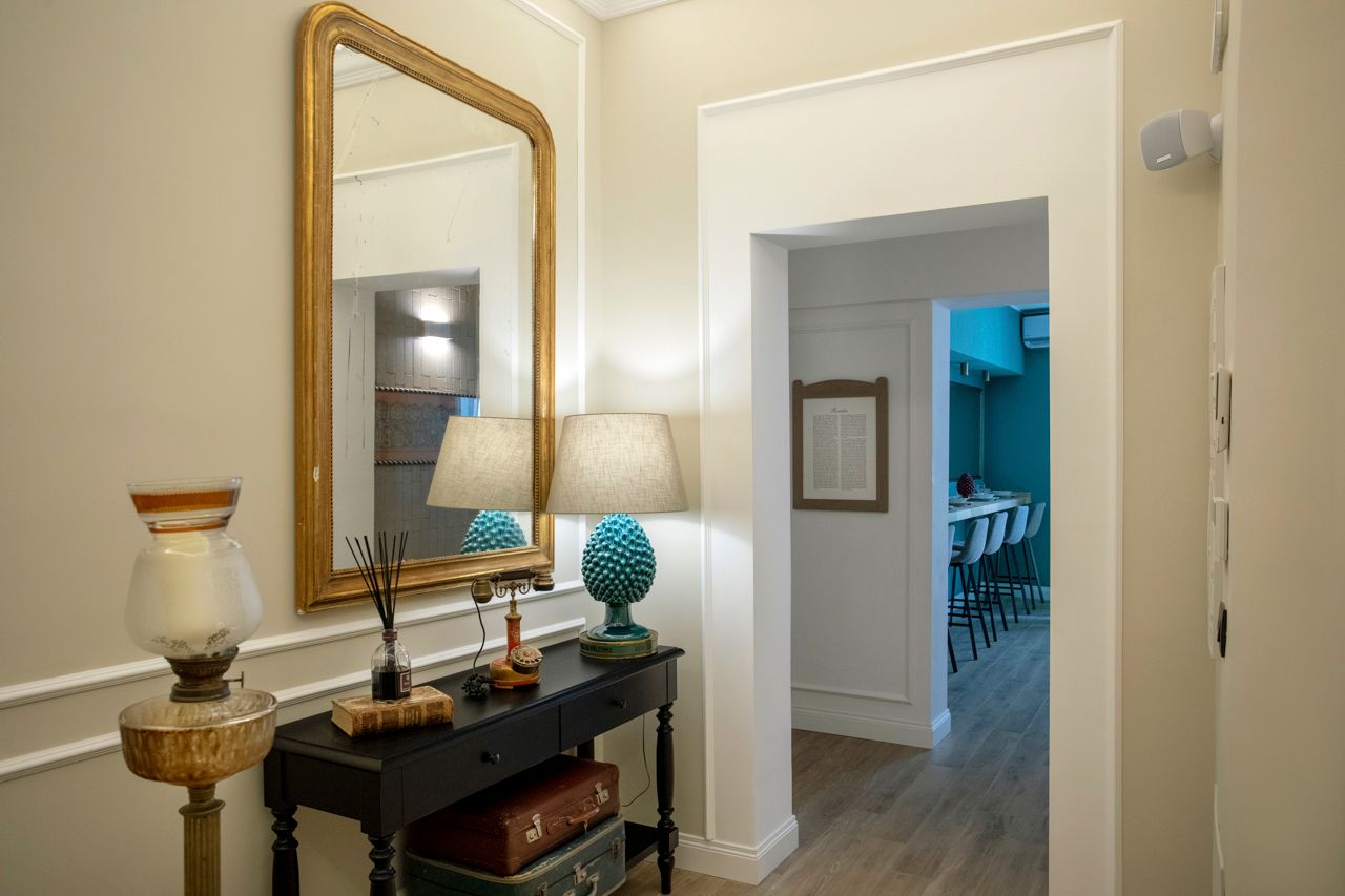 Corridoio con specchio con cornice dorata, consolle e porta che conduce a una stanza color verde acqua.