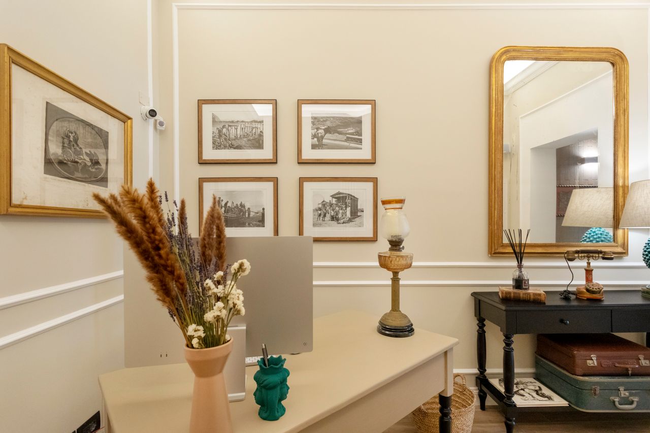 Zona scrivania con opere d'arte incorniciate, uno specchio, una lampada e fiori secchi.
