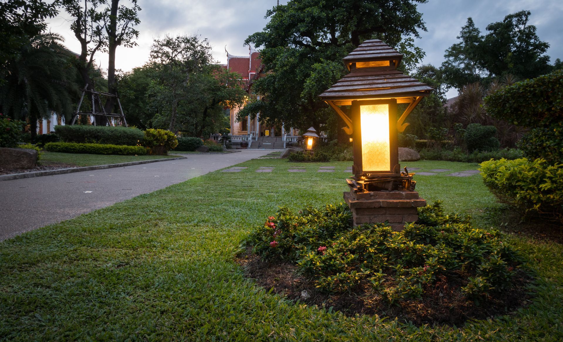 Lit garden lantern beside a grassy pathway at dusk, surrounded by trees and shrubs.