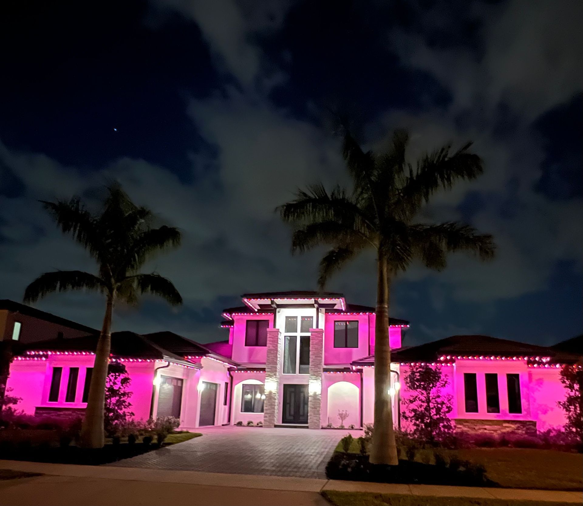 House illuminated in pink with palm trees, under a night sky with clouds.