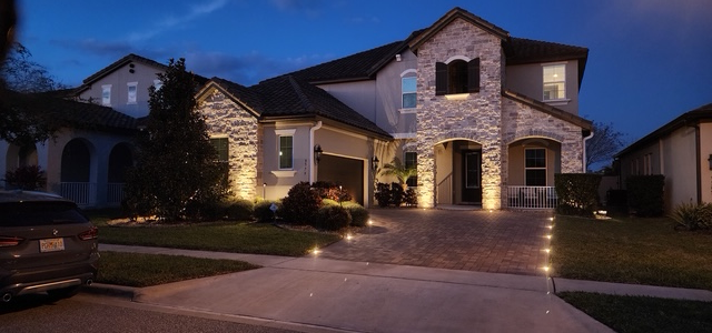 A two-story stone house at dusk, illuminated by glowing landscaping lights along the driveway and facade.