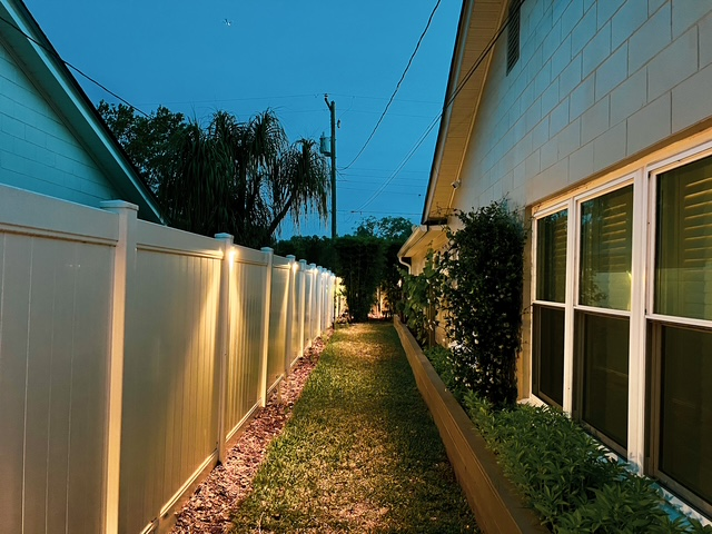 A narrow, grassy pathway between a white vinyl fence illuminated by wall lights and the side of a house at twilight.