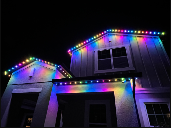 A house at night illuminated by colorful, rainbow-toned LED string lights outlining the rooflines and entryway.