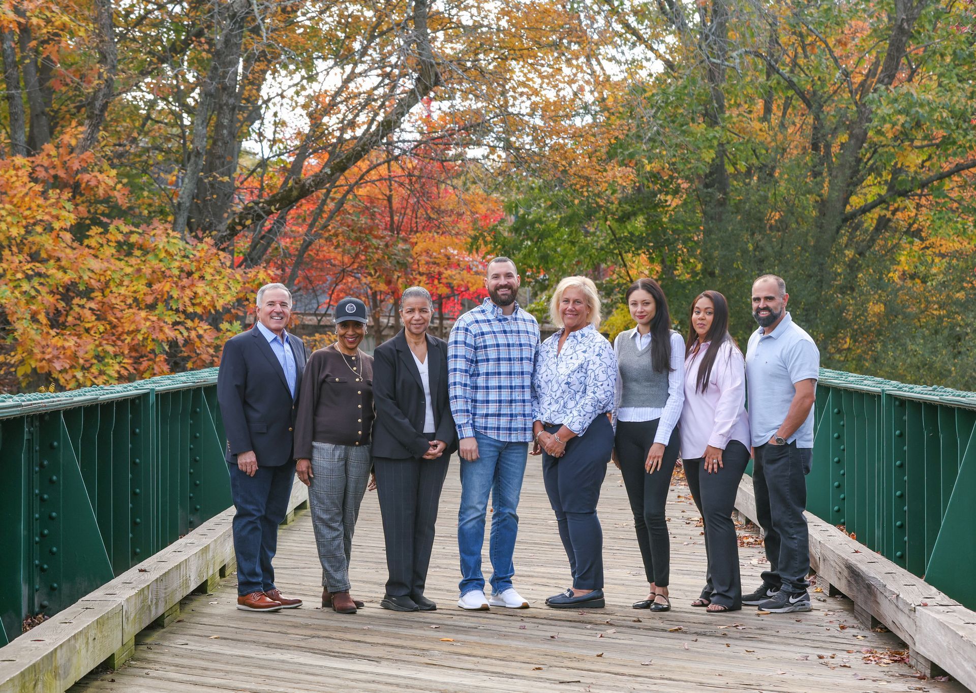 Group of eight people standing on a green bridge with autumn trees in the background.