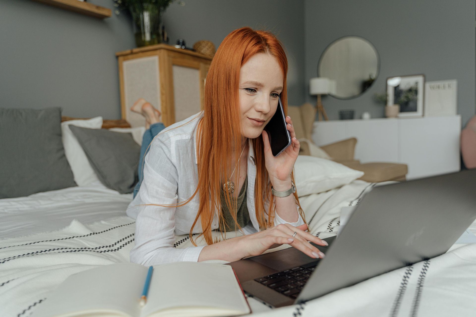 Woman with red hair on phone, using laptop while lying on a bed, notepad open.