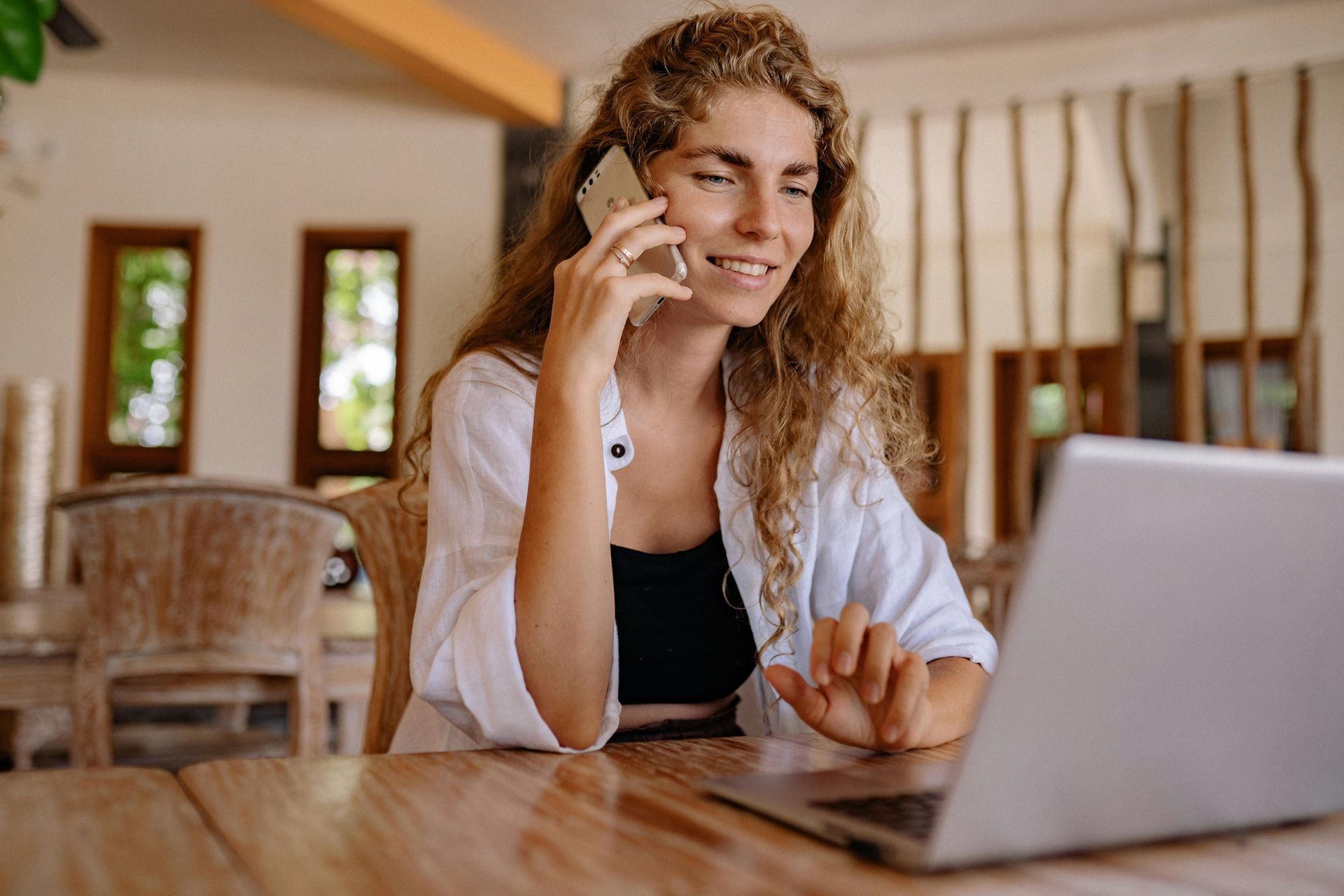 Woman on phone smiles while working on laptop at a table.