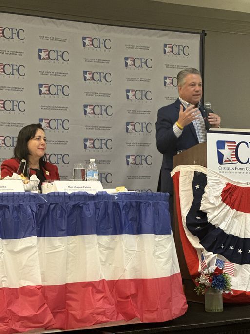 A man stands at a podium giving a speech while a woman sits at a table