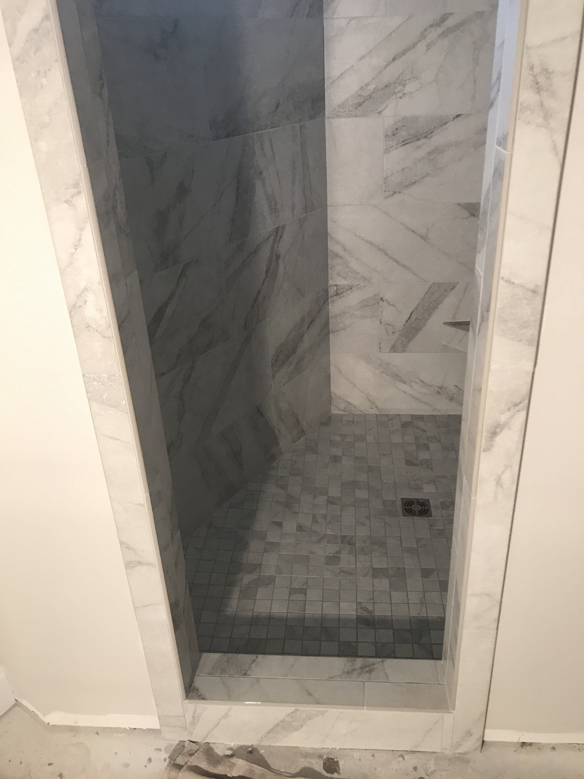 Marble-tiled shower niche with a gray stone mosaic floor, framed by white trim against a white wall.
