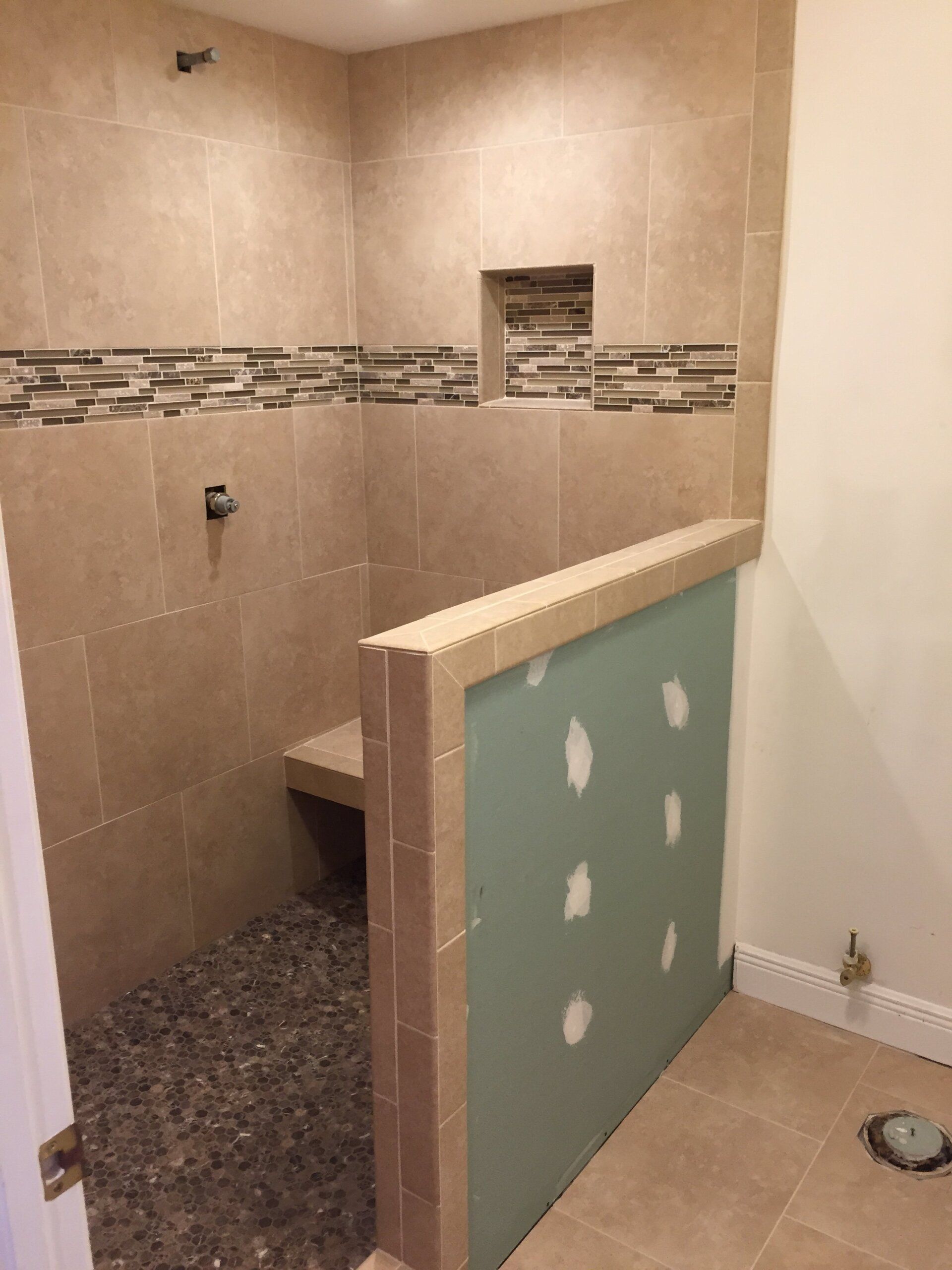 Shower stall with tan tile walls, pebble floor, built-in seat, and green drywall section.