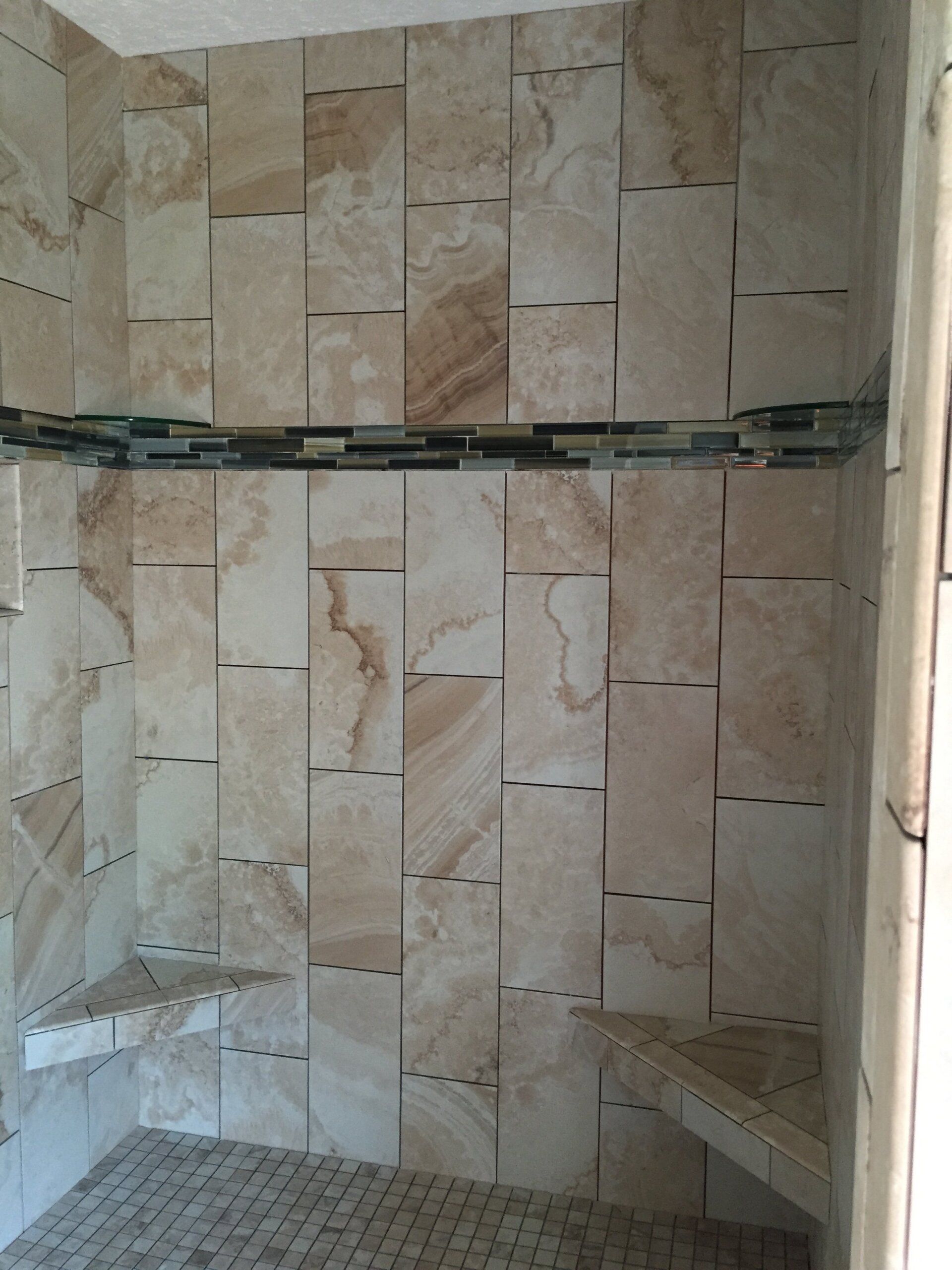 Shower with beige stone tiles, two corner shelves, and mosaic floor.