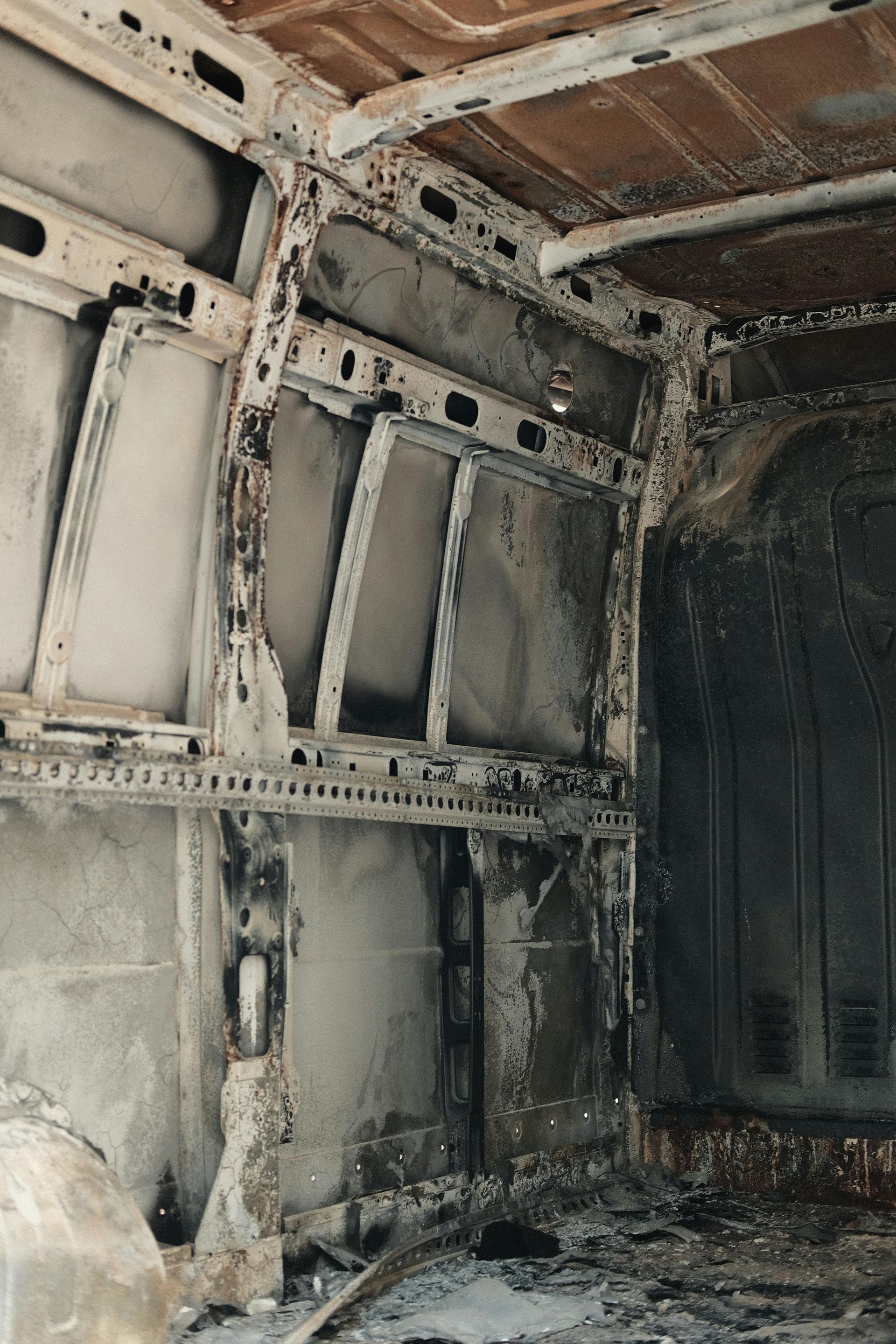Interior of a charred van, revealing metal framework and blackened walls.