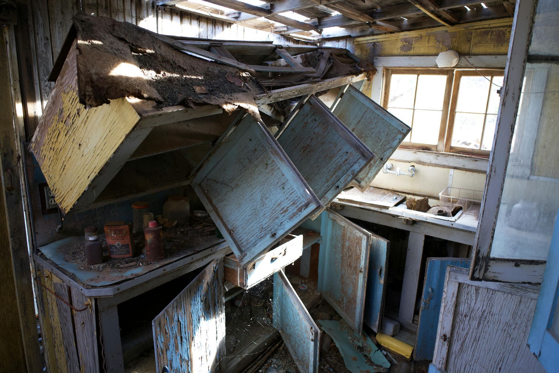 Collapsed kitchen with falling cabinets and debris; sunlight streams through windows.