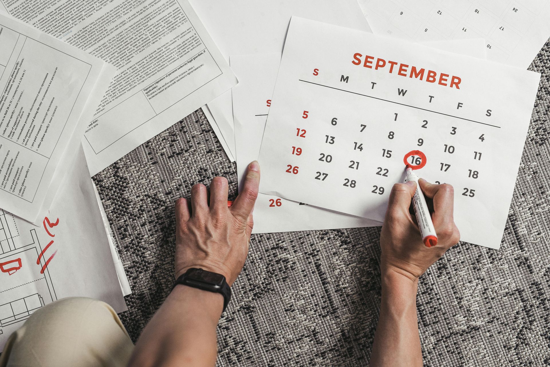 Hands circling a date on a September calendar with a red marker, surrounded by documents on a carpet.