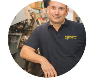 Een man in een blauw shirt met een logo erop, leunend tegen een machine in een werkplaats.
