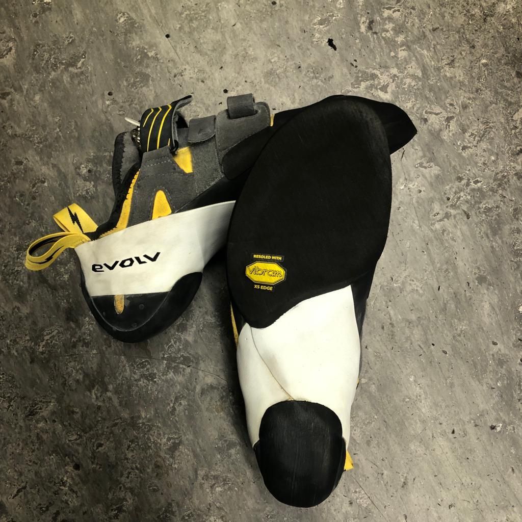 Een paar zwarte, gele en witte Evolv-klimschoenen op een grijs oppervlak.