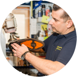 Schoenmaker werkt in zijn werkplaats aan een schoen, met behulp van machines. Hij draagt ​​een blauw shirt en horloge.