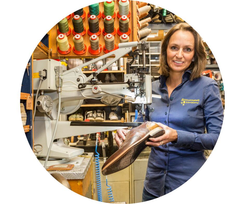 Vrouw in een werkplaats, met een schoen in haar hand. Ze glimlacht, met machines en klosjes garen op de achtergrond.