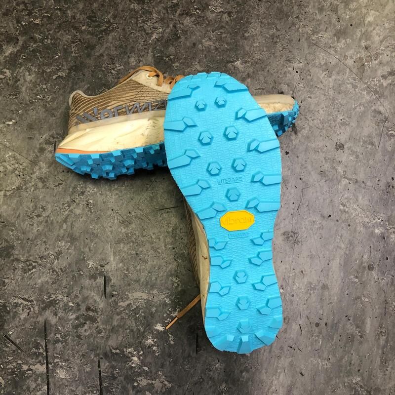 Bruine trailschoen met lichtblauwe Vibram-zool, met een profiel op een grijs oppervlak.
