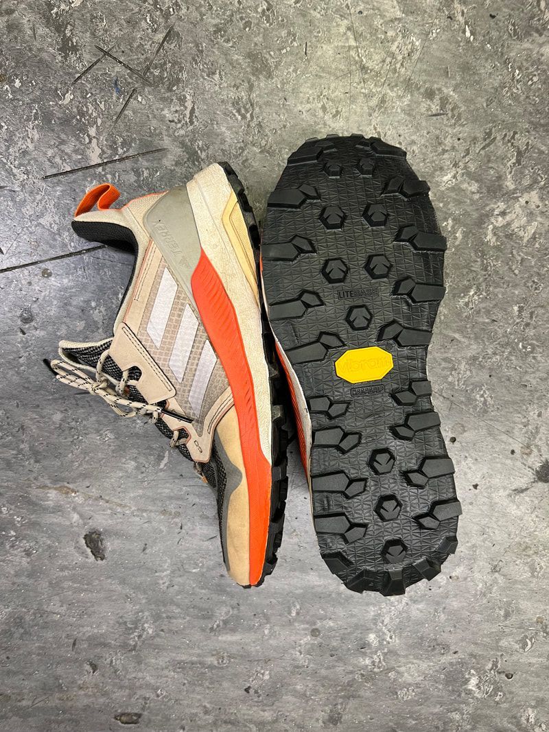 Een bruine en oranje Adidas-wandelschoen met een zichtbare Vibram-zool, liggend op een betonnen ondergrond.