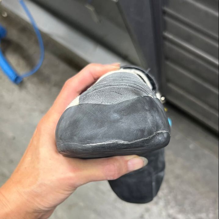 De hand van de persoon houdt een versleten klimschoen vast, waarvan de neus en zool zichtbaar zijn. Zwart rubber en lichte slijtageplekken zijn zichtbaar.