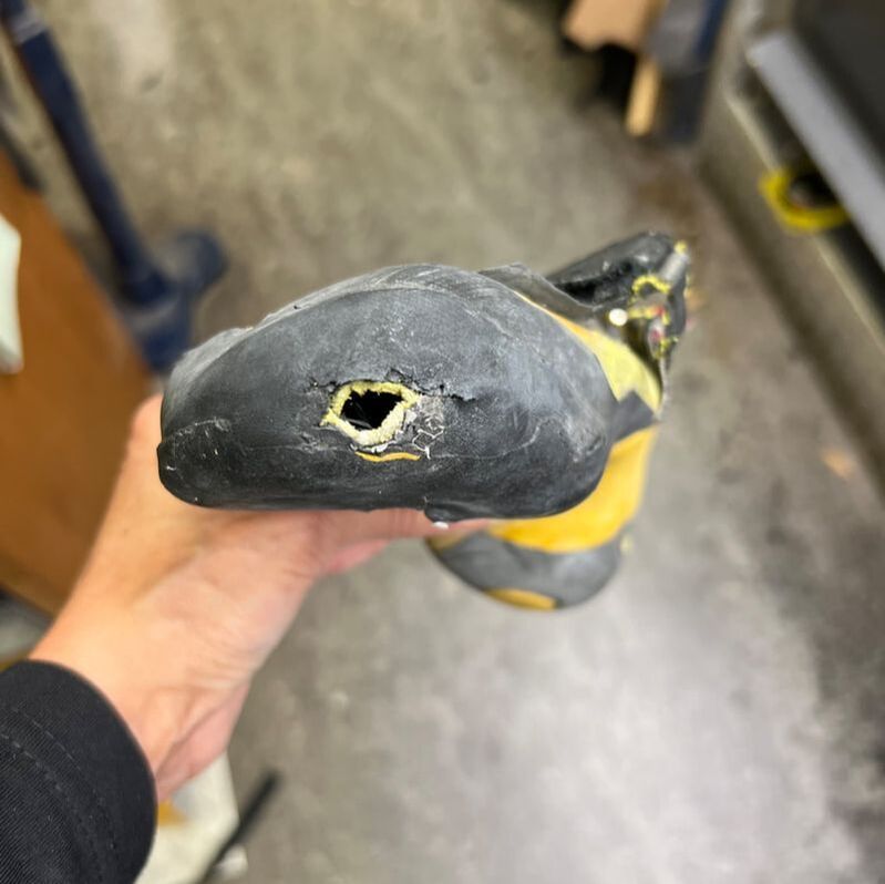 Hand die een versleten klimschoen vasthoudt met een gat in de rubberen neus. De kleuren zwart, geel en grijs zijn zichtbaar.