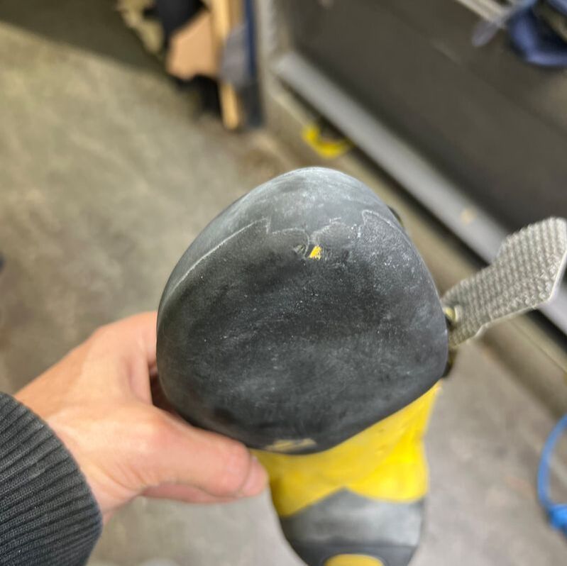 In zijn hand houdt hij een zwart-gele klimschoen vast, waarvan de neus slijtage vertoont.