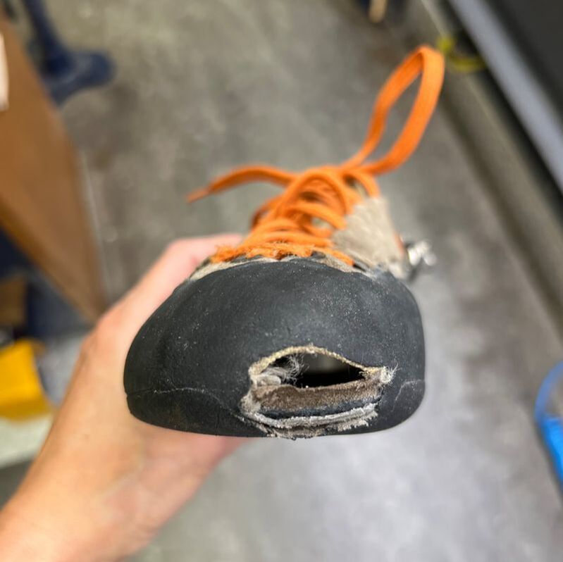 Versleten klimschoen met een groot gat in de neus. Oranje veters, zwarte rubberen zool.