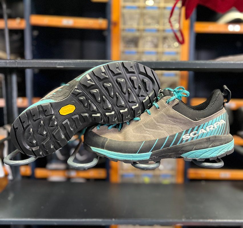 Een paar Scarpa wandelschoenen met een grijze bovenkant, blauwgroene accenten en zwarte zolen. Eén schoen staat ondersteboven.