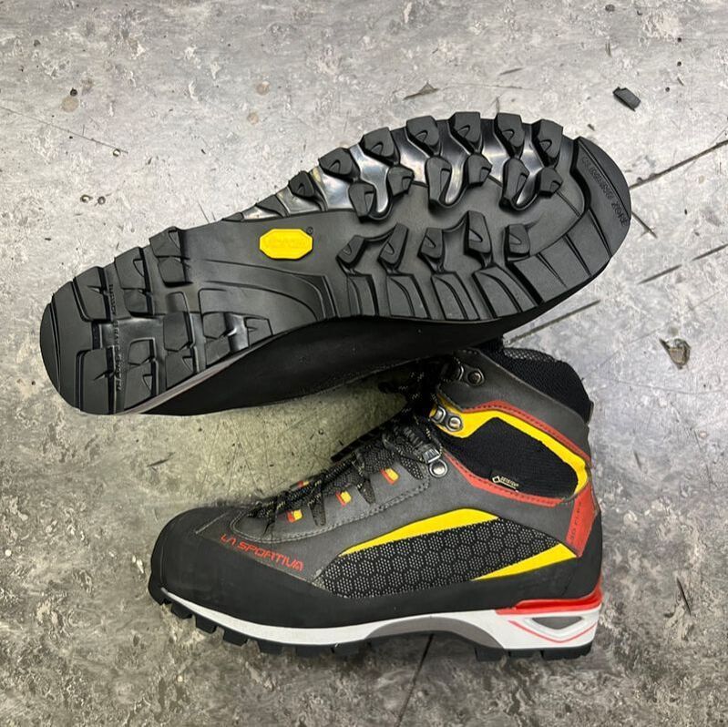 Een paar wandelschoenen met een geribbelde zool, grijs bovenwerk met gele en rode accenten, rustend op beton.