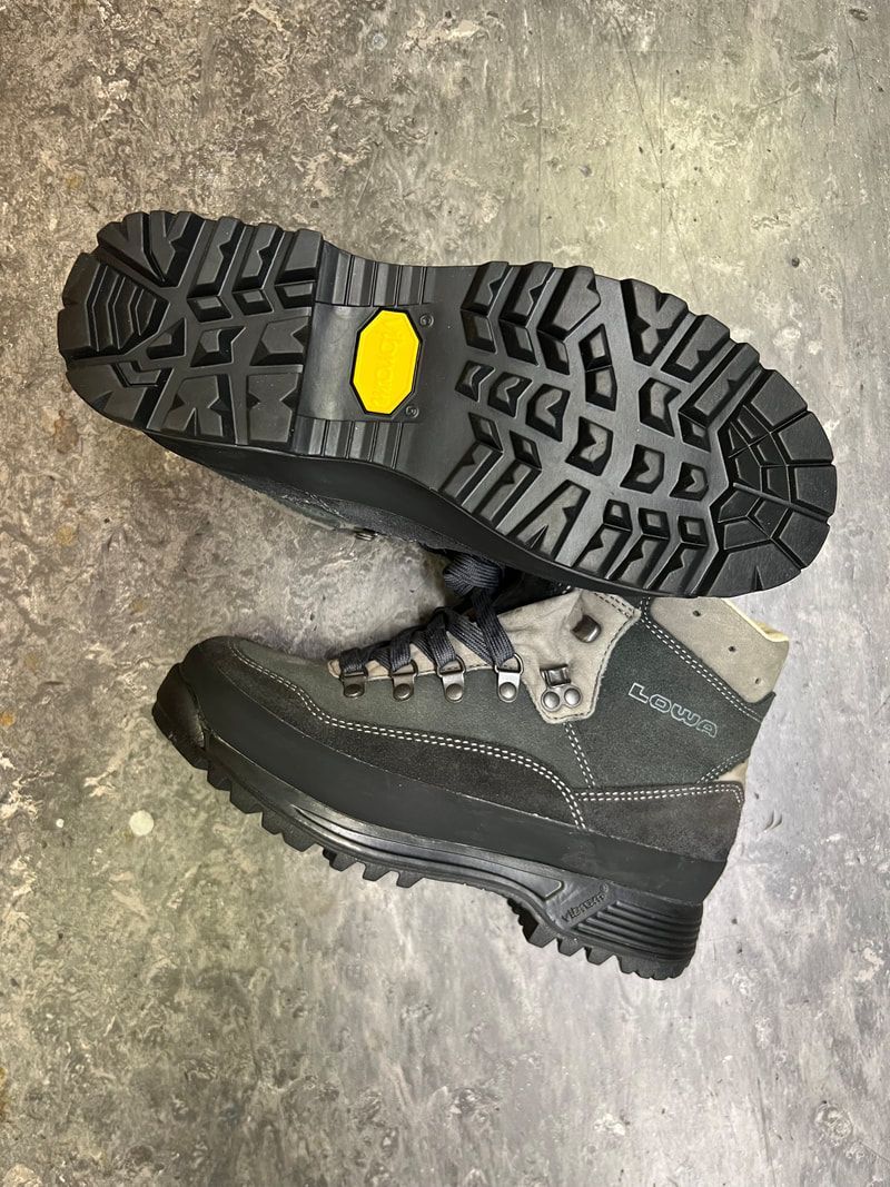 Wandelschoenen: zwarte, geribbelde zool met geel Vibram-logo. Bovenwerk van grijs suède en zwart leer, veters zichtbaar.