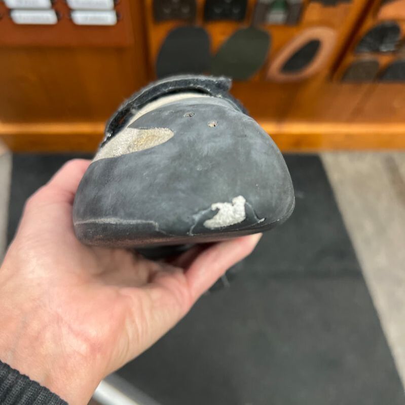 Hand die een versleten, donkere klimschoen vasthoudt, waarvan de neus beschadigd is.