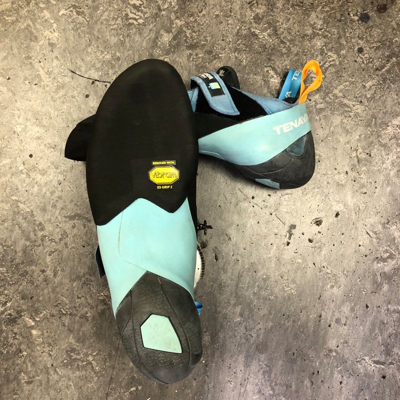 Een paar turquoise en zwarte Tenaya-klimschoenen op een grijze ondergrond. Eén schoen staat ondersteboven.