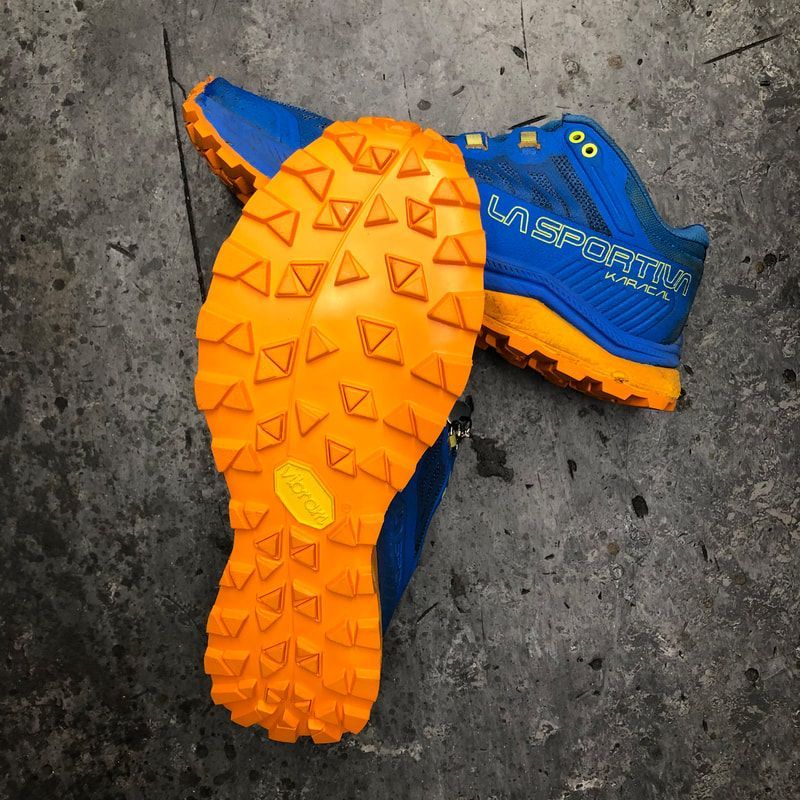 Blauwe en oranje La Sportiva trailschoenen op een grijze ondergrond, met een opvallende oranje Vibram-zool.