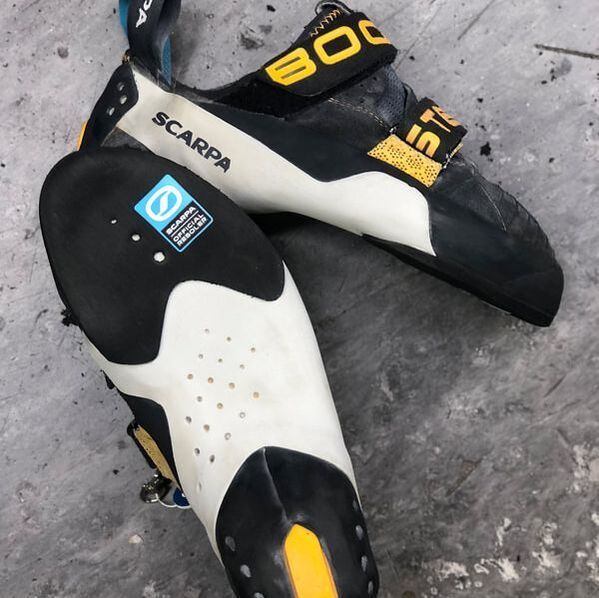 Een paar Scarpa-klimschoenen op beton. Witte, zwarte en gele accenten zijn zichtbaar.