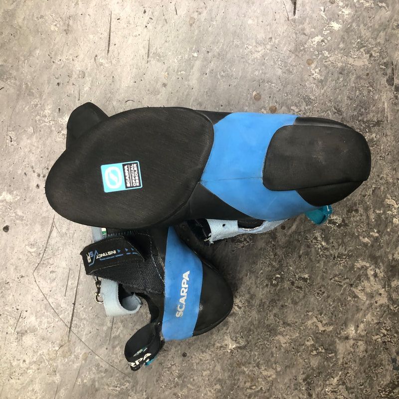 Een paar Scarpa-klimschoenen op een betonnen vloer. Zwarte rubberen zolen met blauwe accenten.