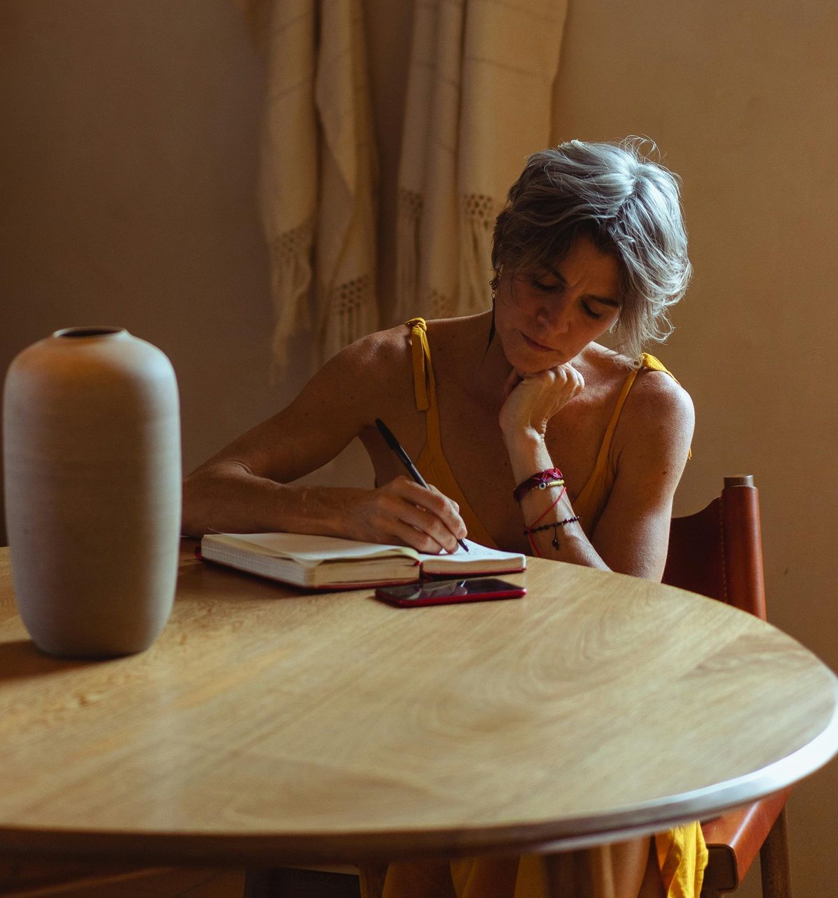older-woman-sitting-at-dining-table-with-vase-contemplating-and-writing-her-life-story-with-pen-and-notebook