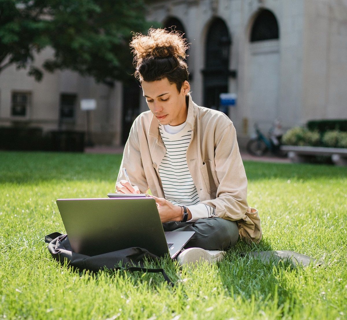 young-man-contemplating-writing-eulogy-on-laptop-sitting-on-grass-in-park-with-large-white-building-behind