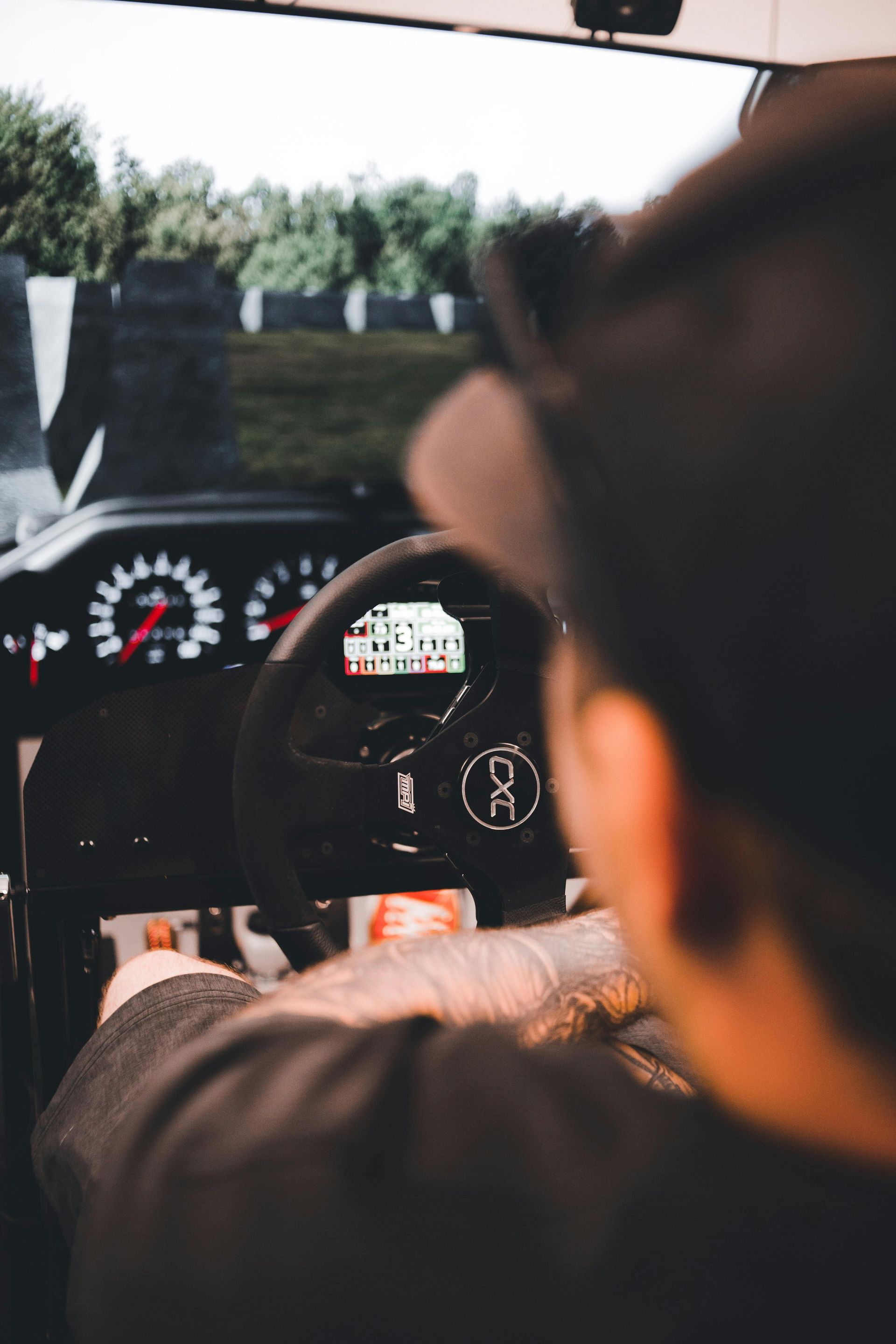 Person using a racing simulator, focused on the steering wheel with digital dashboard, track visible in the background.