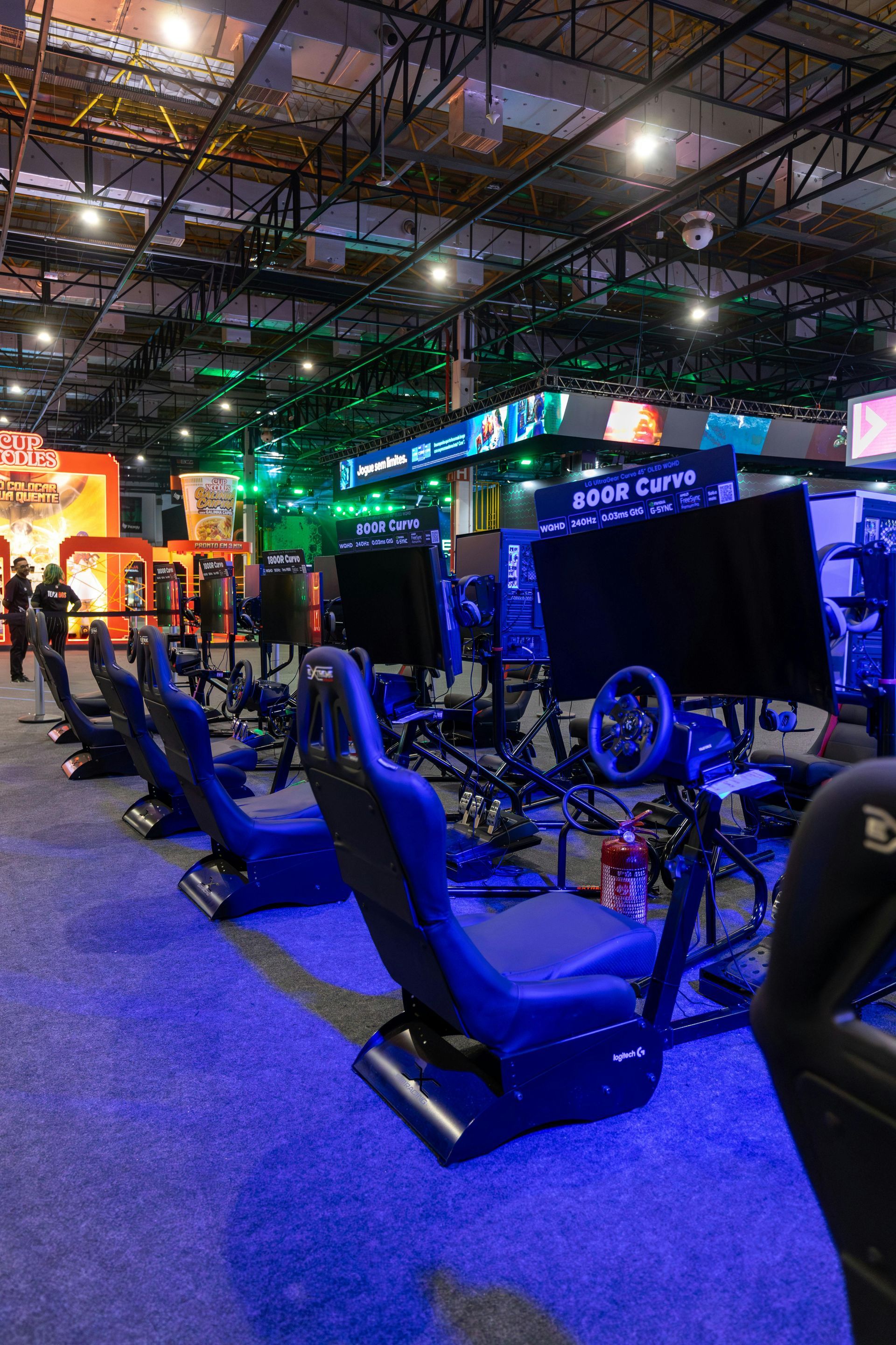Racing game simulators at a gaming event. Blue chairs, black screens, and steering wheels in a large hall.