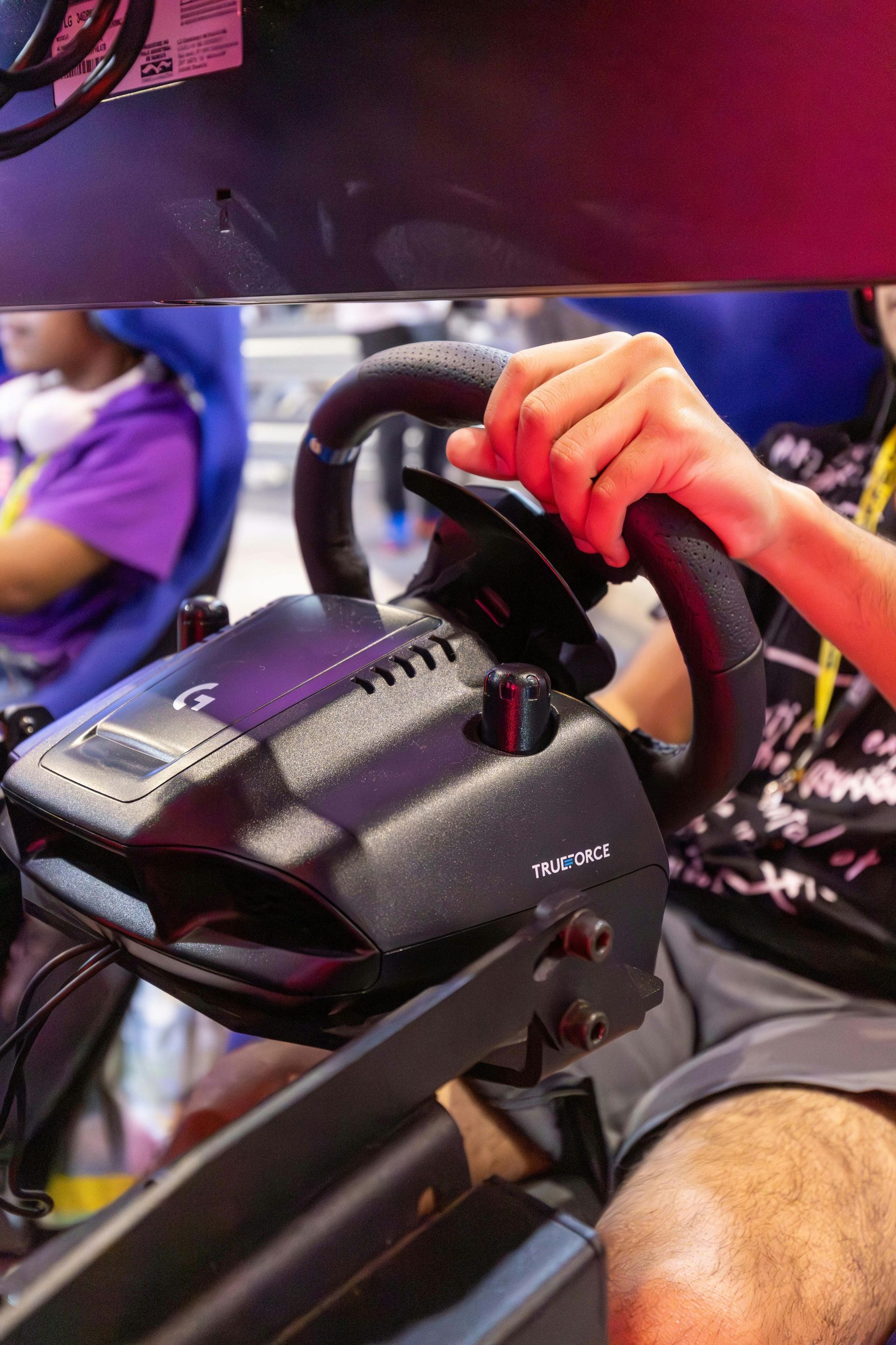Person's hand gripping a black racing wheel with buttons. The wheel is mounted on a black stand.