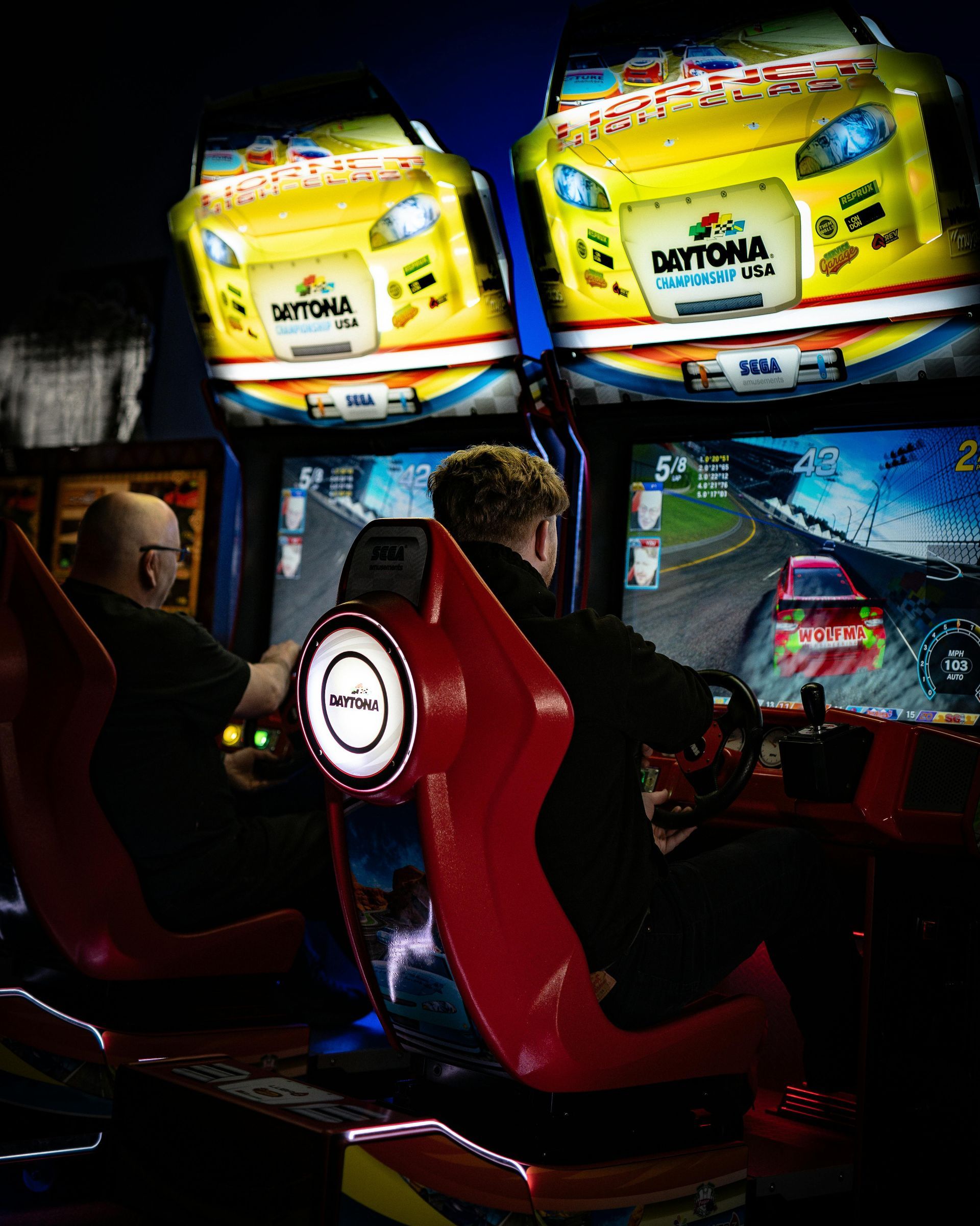 Two people playing Daytona racing arcade games in a dimly lit arcade.
