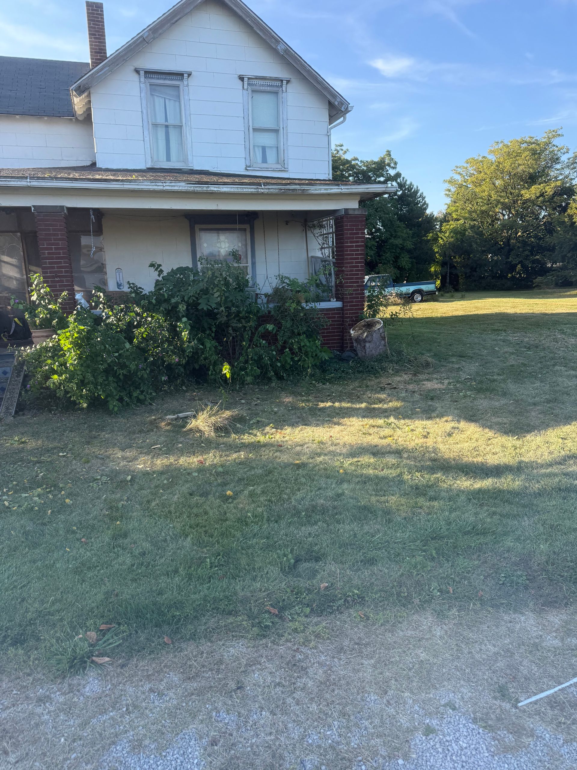 Old, white two-story house with overgrown bushes and a porch. Green lawn in front, blue sky.