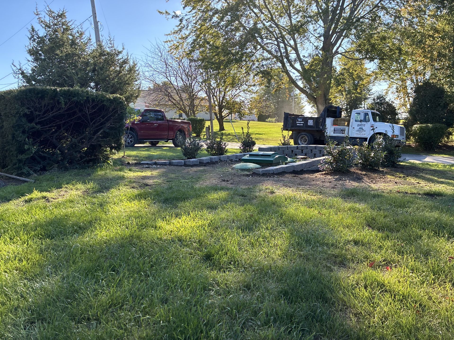 Lawn with landscaping materials, a red pickup truck, and a white dump truck, on a sunny day.