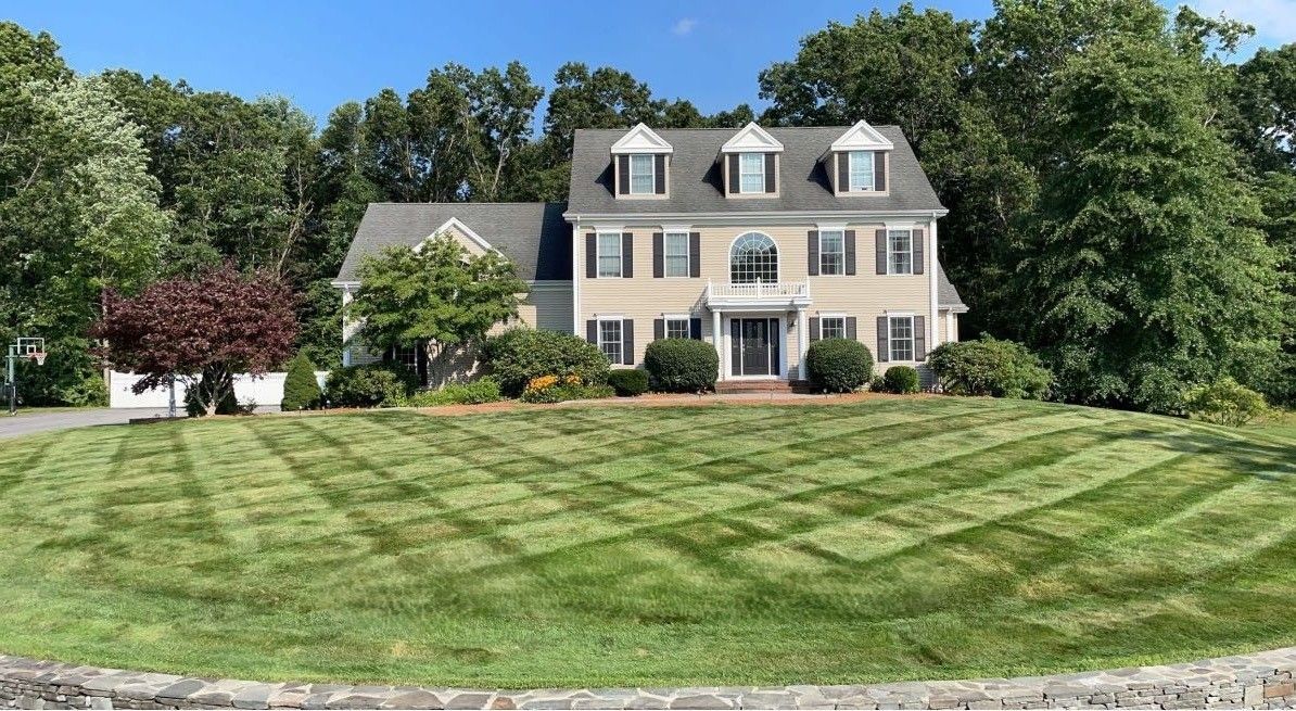 Two-story beige house with manicured lawn featuring a diamond-patterned mowing design. Green trees surround the house.