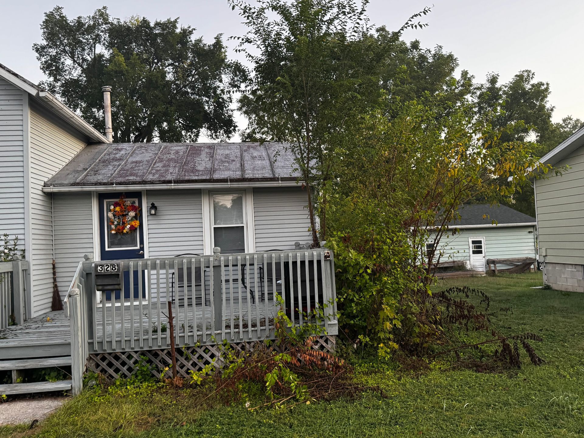 Small, weathered gray house with a wooden deck and overgrown bushes; another small building is visible in the background.