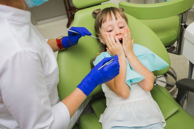 A worried looking little girl is sitting in a dental chair while a dentist examines her teeth.