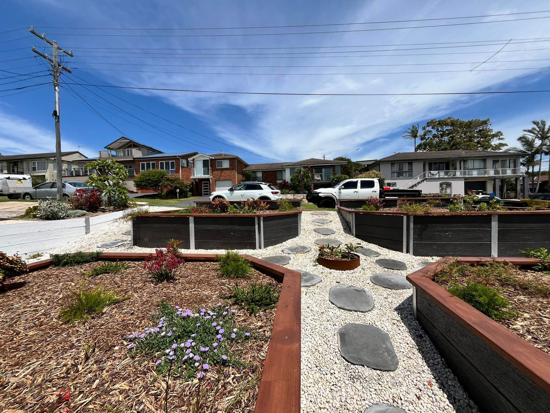 A front yard with raised garden beds and stone pathway. Cars parked on the street, houses in the background.  — CAW Services in Badagarang, NSWBlue sky.
